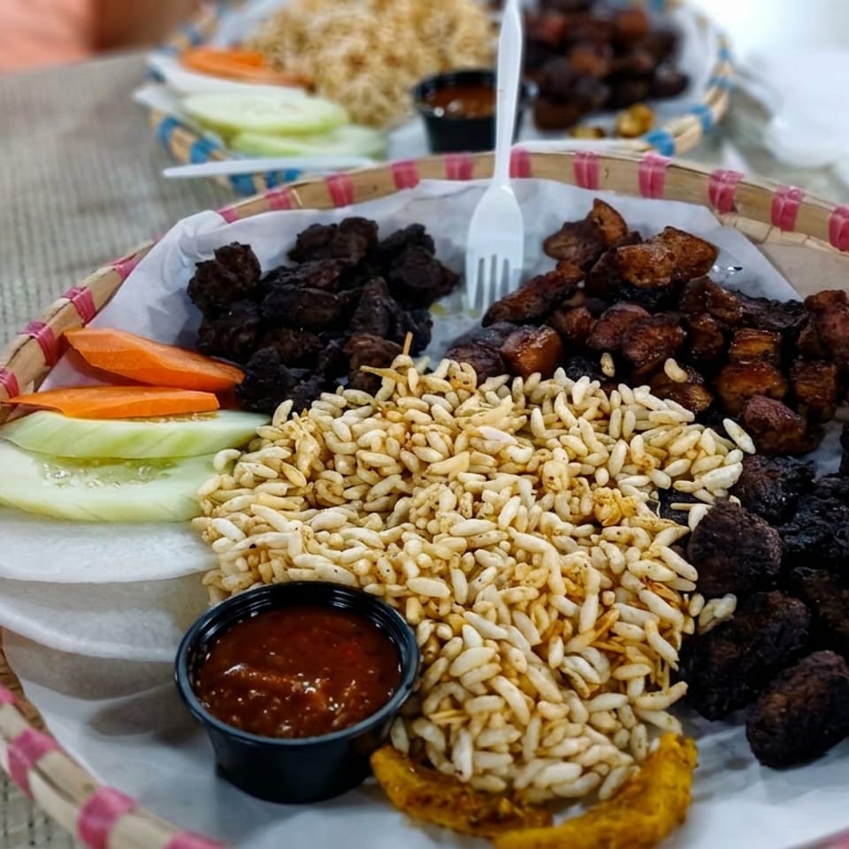 Bamboo tray of grilled meat, puffed rice, sliced carrots and cucumbers, dipping sauce, and fried plantain.