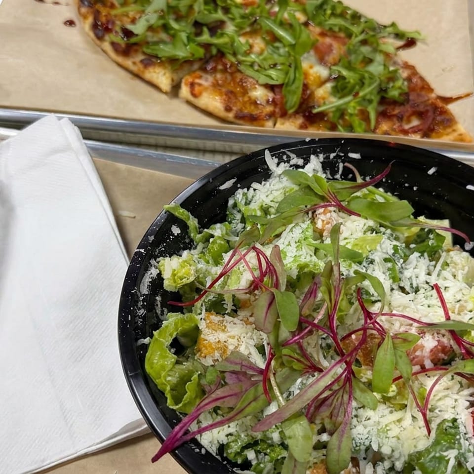 Fresh salad with lettuce, shredded cheese, tomatoes, and microgreens beside a flatbread pizza on cafeteria trays.