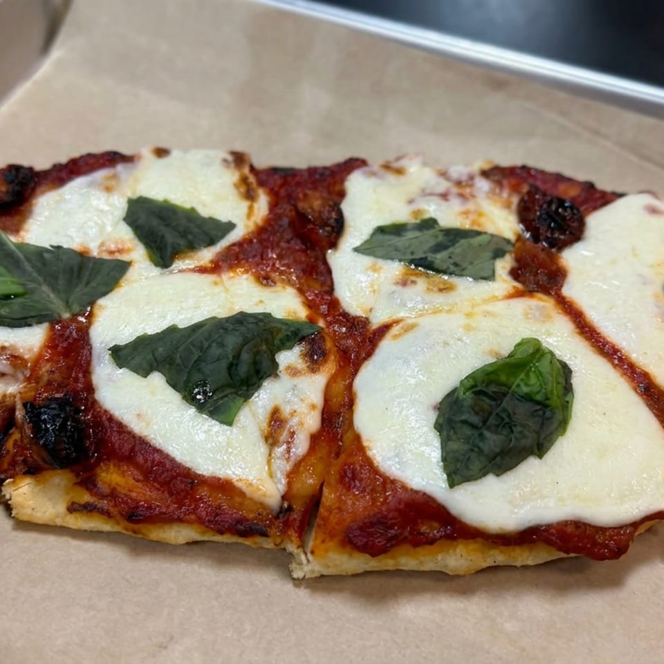 Rectangular pizza topped with tomato sauce, melted mozzarella, fresh basil leaves, and charred olives on a paper-lined tray.